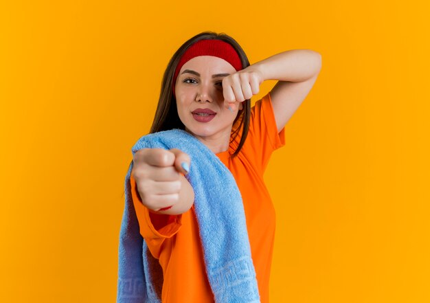 Mulher jovem e confiante desportiva usando bandana e pulseiras com toalha e no ombro fazendo gesto de boxe isolado na parede laranja com espaço de cópia