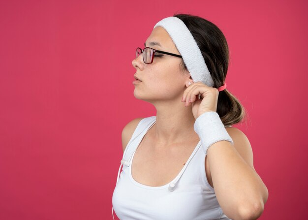Mulher jovem e confiante, esportiva, usando óculos ópticos, fones de ouvido, bandana e pulseira, segurando a orelha e olhando para o lado isolado na parede rosa