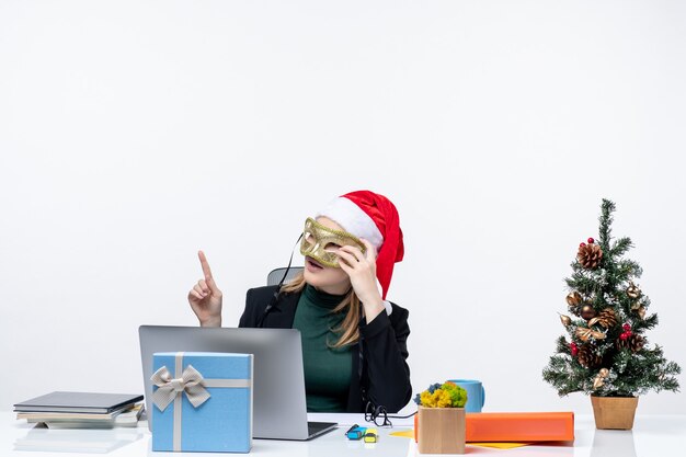 Mulher jovem e curiosa com chapéu de Papai Noel segurando óculos e usando máscara, sentada em uma mesa com uma árvore de Natal e um presente no escritório em fundo branco