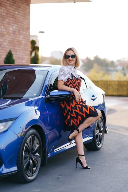 Mulher jovem e deslumbrante com um vestido posando na frente de seu carro ao ar livre.