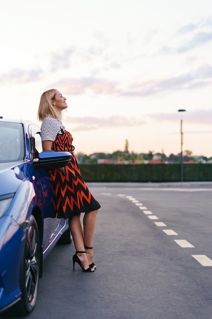 Foto grátis mulher jovem e deslumbrante com um vestido posando na frente de seu carro na estrada com o céu ao fundo.