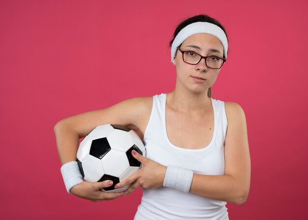 Mulher jovem e desportiva desapontada com óculos ópticos, fita para a cabeça e pulseira, segurando uma bola isolada na parede rosa