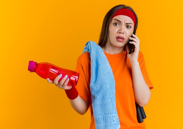 Mulher jovem e desportiva insatisfeita usando bandana e pulseiras com corda de pular e toalha nos ombros, segurando uma garrafa de água e falando no telefone, olhando para o lado