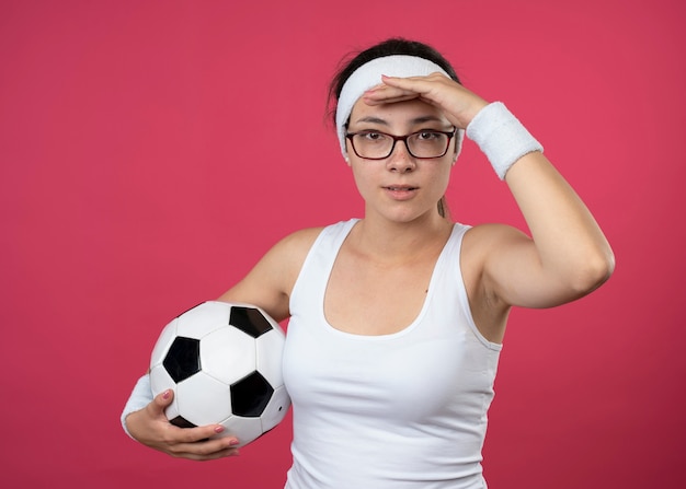 Foto grátis mulher jovem e desportiva satisfeita com óculos ópticos, bandana e pulseira, mantém a palma da mão na testa e segura a bola isolada na parede rosa