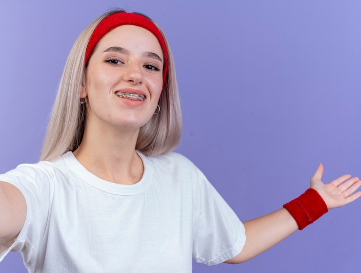 Foto grátis mulher jovem e desportiva sorridente com aparelho usando fita para a cabeça e pulseiras com a mão aberta e olhando para a frente, isolada na parede roxa