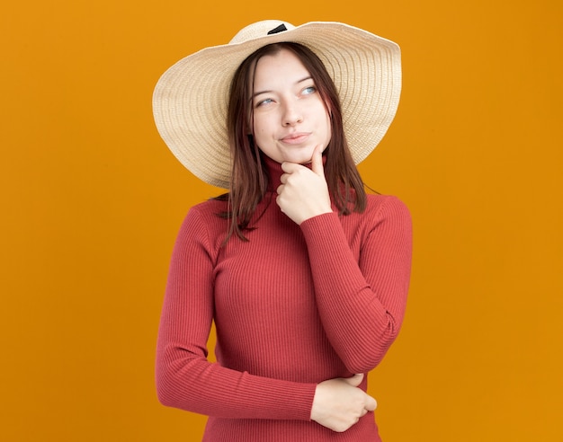 Mulher jovem e duvidosa com chapéu de praia, segurando o queixo, olhando para o lado isolado na parede laranja