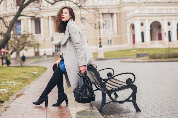 Mulher jovem e elegante caminhando na cidade de outono, estação fria, usando botas pretas de salto alto, mochila de couro, acessórios, casaco cinza, sentada no banco, tendência da moda