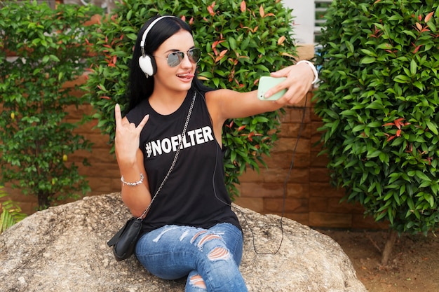 Mulher jovem e elegante hippie em camiseta preta, jeans, ouvindo música em fones de ouvido, se divertindo, posando, tirando foto de selfie no telefone, mostrando o símbolo da paz, expressão de carinha engraçada