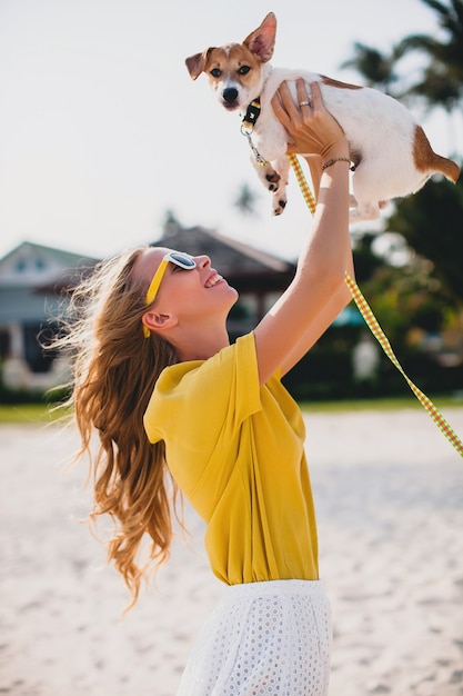 Foto grátis mulher jovem e elegante hippie segurando andando brincando de cachorro cachorrinho jack russell, parque tropical, sorrindo e se divertindo, férias, óculos de sol, boné, camisa amarela, areia da praia