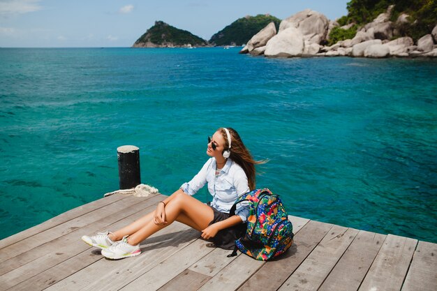 Mulher jovem e elegante hippie viajando ao redor do mundo, sentada no cais, óculos de aviador, fones de ouvido, ouvindo música, férias, mochila, camisa jeans, feliz, lagoa de ilha tropical