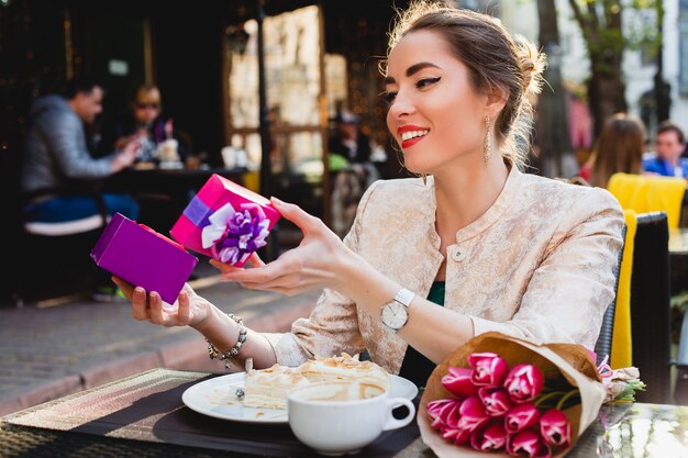 Mulher jovem e elegante sentada em um café, segurando uma caixa de presente, sorrindo