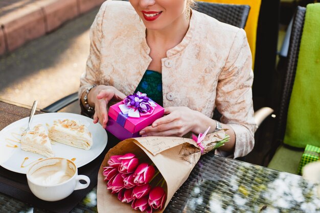 Mulher jovem e elegante sentada em um café, segurando uma caixa de presente, sorrindo