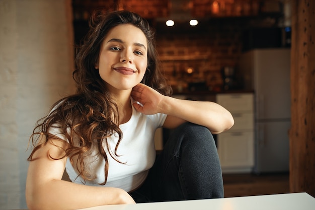 Mulher jovem e encantadora com roupas casuais, relaxando em casa, sentada à mesa com uma cozinha aconchegante no fundo, olhando com um sorriso confiante e feliz. emoções positivas, descanso e relaxamento