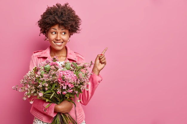 Mulher jovem e encaracolada positiva posa com um lindo buquê de flores apontando para um espaço em branco mostra o conteúdo da publicidade usando uma jaqueta isolada sobre a parede rosa