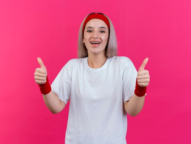 Foto grátis mulher jovem e esportiva empolgada com aparelho usando fita para a cabeça e pulseiras com o polegar para cima de duas mãos isoladas na parede rosa