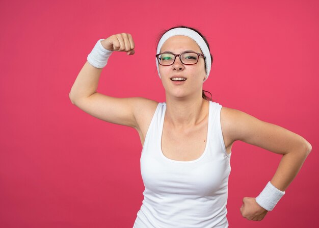 Mulher jovem e esportiva irritada usando óculos ópticos, bandana e pulseira, segurando os punhos para cima e para baixo, isolados na parede rosa