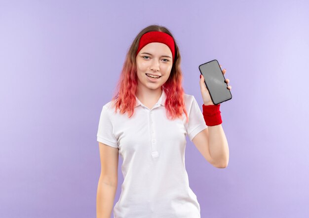 Mulher jovem e esportiva mostrando smartphone sorrindo com uma cara feliz em pé sobre a parede roxa