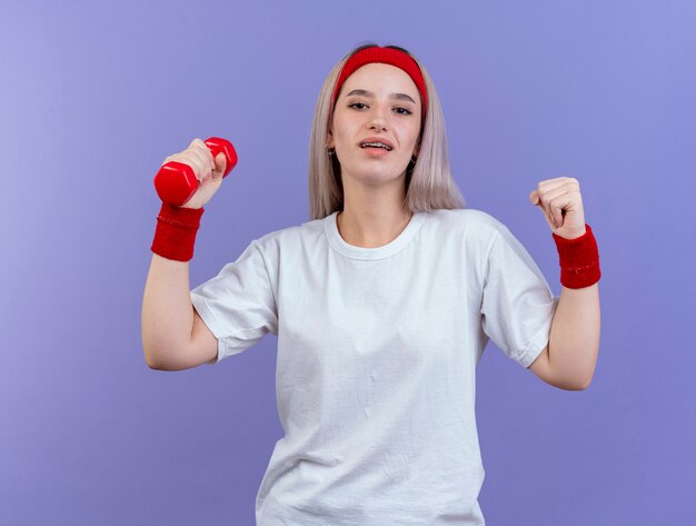 Mulher jovem e esportiva satisfeita com aparelho usando fita para a cabeça e pulseiras, mantendo o punho e segurando halteres isolado na parede roxa