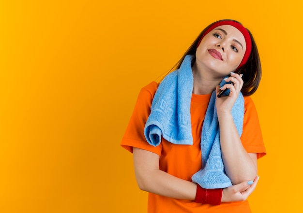 Mulher jovem e esportiva satisfeita usando bandana e pulseiras com uma toalha no pescoço olhando para o lado colocando a mão no cotovelo falando ao telefone