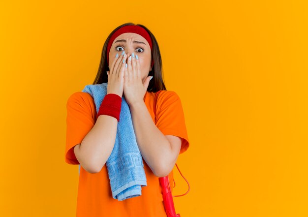 Mulher jovem e esportiva surpresa usando bandana e pulseiras com toalha e pular corda nos ombros, mantendo as mãos na boca isolada na parede laranja com espaço de cópia