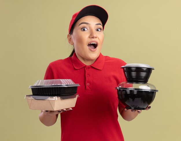 Mulher jovem e feliz entregadora de uniforme vermelho e boné segurando pacotes de comida olhando para a frente feliz e surpresa em pé sobre a parede verde