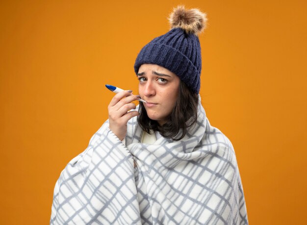 Mulher jovem e fraca com um manto de inverno enrolado em uma manta, colocando o termômetro na boca, olhando para a frente, isolado na parede laranja