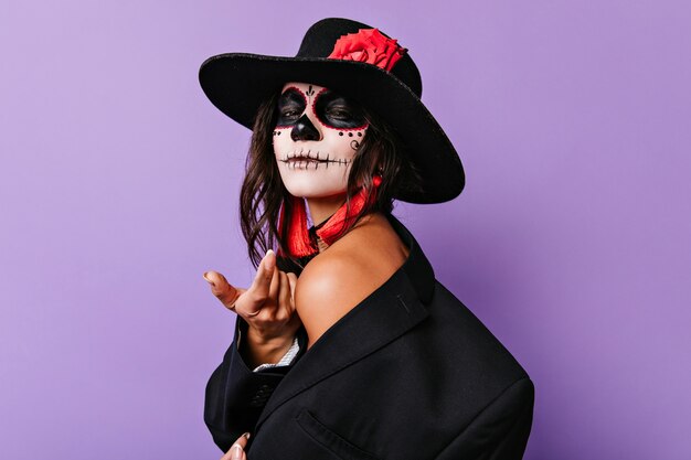 Mulher jovem e graciosa de sombrero preto em pé na parede roxa. Menina morena despreocupada com maquiagem de halloween, sorrindo suavemente.