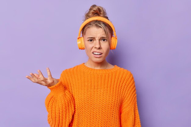 Mulher jovem e indignada e perplexa veste um suéter de malha casual levanta a palma da mão parece sem noção, não consigo ouvir você enquanto ouve música em poses de fones de ouvido contra um fundo roxo. Conceito de reações humanas.