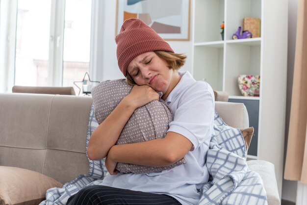 Mulher jovem e insalubre chateada com um chapéu quente com um cobertor parecendo indisposto e doente, sofrendo de resfriado e gripe, segurando o travesseiro com uma expressão triste no rosto, sentada no sofá em uma sala iluminada