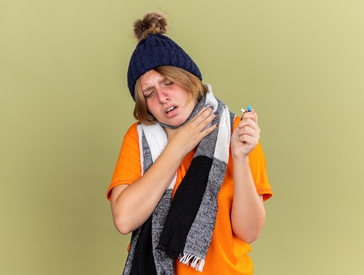 Foto grátis mulher jovem e insalubre com chapéu e lenço no pescoço, sentindo-se mal, segurando vários comprimidos para gripe e dor de garganta, tocando o pescoço em pé sobre a parede verde
