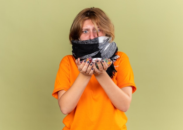 Foto grátis mulher jovem e insalubre em uma camiseta laranja com um lenço quente em volta do pescoço sentindo uma terrível gripe, segurando vários comprimidos e parecendo preocupada