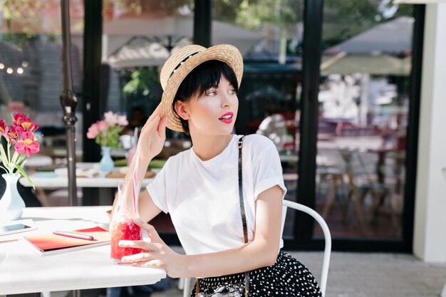 Mulher jovem e linda com um penteado da moda relaxando em um restaurante ao ar livre e olhando para longe enquanto bebe um coquetel