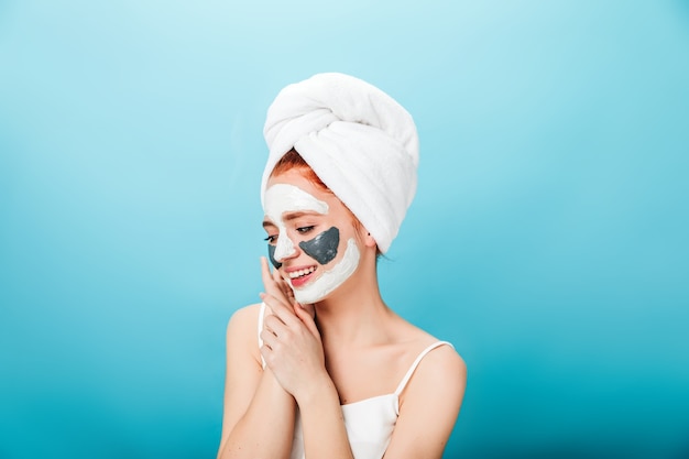 Mulher jovem e linda em toalha posando sobre fundo azul. Foto de estúdio de menina branca com máscara facial.