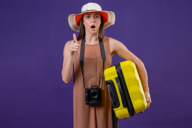 Mulher jovem e linda viajante com chapéu de verão com mala amarela e câmera fotográfica apontando com o dedo para a câmera, surpresa e maravilhada de pé sobre o fundo roxo