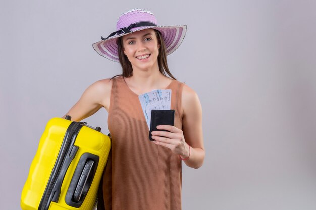 Mulher jovem e linda viajante com chapéu de verão segurando uma mala amarela e bilhetes sorrindo com uma cara feliz em pé sobre um fundo branco