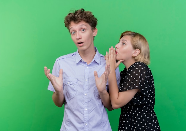 Foto grátis mulher jovem e lindo casal sussurrando um segredo para o namorado surpreso em pé sobre a parede verde