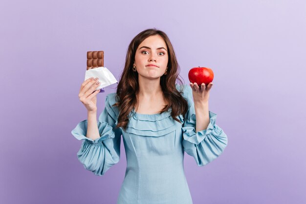 Mulher jovem e magra escolhe entre maçã saudável e chocolate doce. A morena não consegue decidir o que comer no almoço.