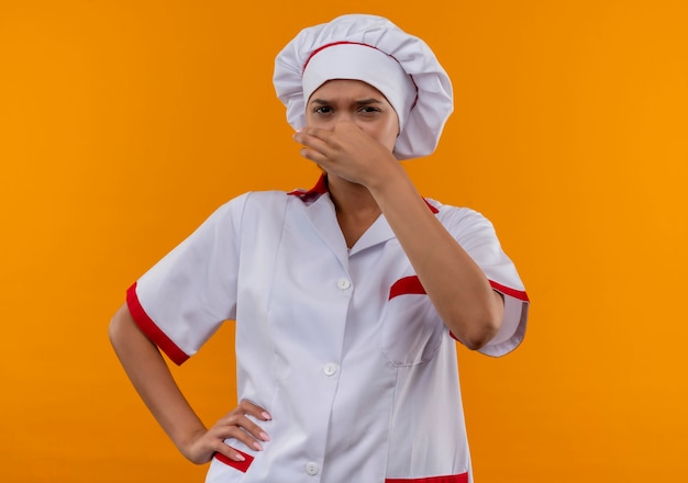 Mulher jovem e melindrosa, cozinheira, vestindo uniforme de chef, fechou o nariz, colocando a mão no quadril em um fundo laranja isolado