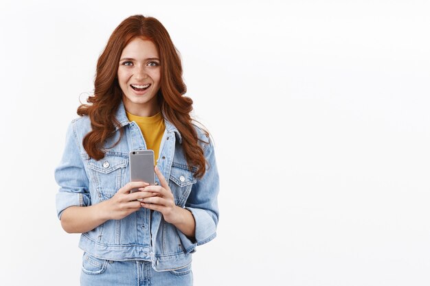 Mulher jovem e moderna alegre em jaqueta jeans, segurando smartphone e sorrindo otimista, tirando fotos, telefone novo, mensagens, parede branca em pé