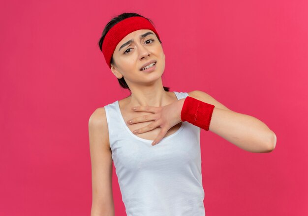 Mulher jovem e preocupada com fitness em roupas esportivas com fita para a cabeça e rosto infeliz, segurando a palma da mão no peito em pé sobre a parede rosa