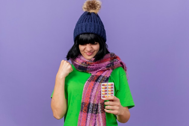 Mulher jovem e satisfeita com um chapéu de inverno e lenço segurando um pacote de cápsulas fazendo o gesto de sim isolado na parede roxa com espaço de cópia