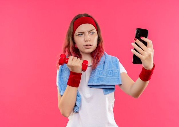 Mulher jovem e séria desportiva com bandana e toalha no ombro tirando uma selfie mostrando halteres na mão para a câmera de seu smartphone em pé sobre a parede rosa