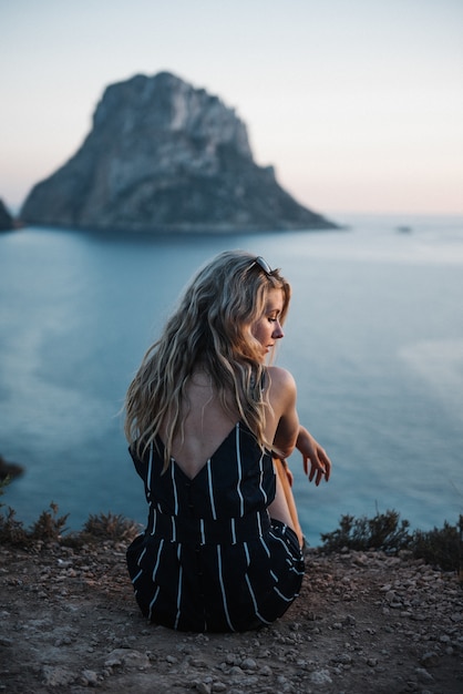 Foto grátis mulher jovem e solitária com cabelo loiro, sentada à beira-mar, aproveitando seu tempo de paz