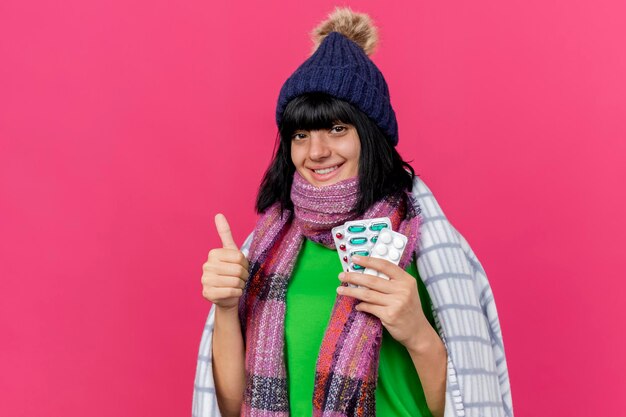 Mulher jovem e sorridente com chapéu de inverno e lenço enrolado em uma manta segurando comprimidos médicos, olhando para a frente, mostrando o polegar isolado na parede rosa com espaço de cópia