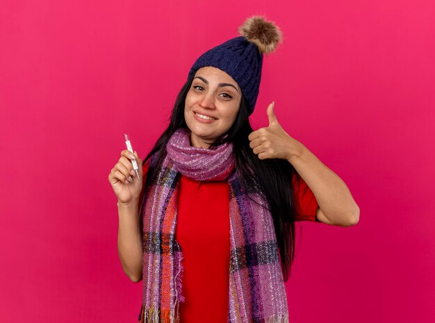 Mulher jovem e sorridente com chapéu de inverno e lenço segurando o termômetro, olhando para frente, mostrando o polegar isolado na parede rosa com espaço de cópia