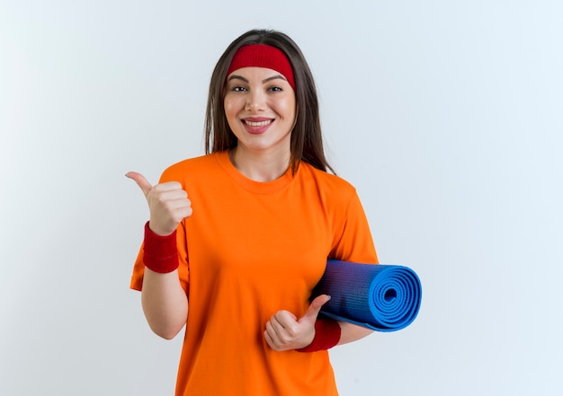 Mulher jovem e sorridente e esportiva usando bandana e pulseiras segurando um tapete de ioga mostrando os polegares isolados na parede branca com espaço de cópia