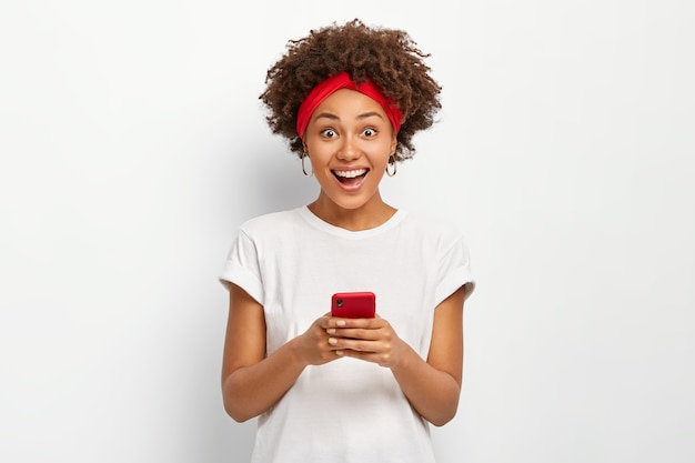 Mulher jovem e sorridente feliz segura um celular moderno, sorri positivamente, navega na internet, cria seu próprio blog, usa uma camiseta branca