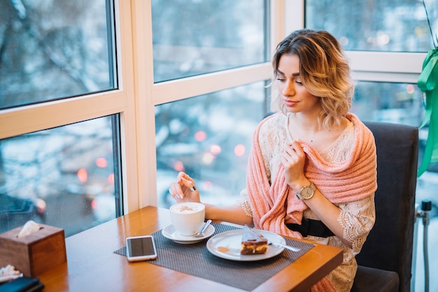 Mulher jovem elegante com sobremesa e copo de bebida perto de smartphone na mesa no café