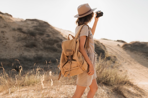 Mulher jovem elegante com vestido cáqui caminhando pelo deserto, viajando pela África em um safári, usando chapéu e mochila, tirando foto com uma câmera vintage