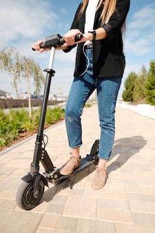 Mulher jovem elegante em roupas casuais inteligentes em pé na scooter elétrica na estrada em ambiente urbano contra árvores verdes e céu nublado